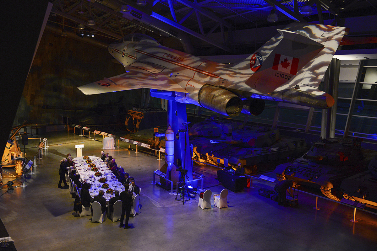 A group of people are sitting around a table in the Canadian War Museum, in Ottawa, with a fighter jet.